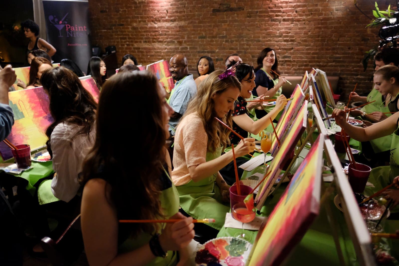 A work team at a corporate painting team building event — each person at an easel painting the same scene while a Paint Nite artist guides the room
