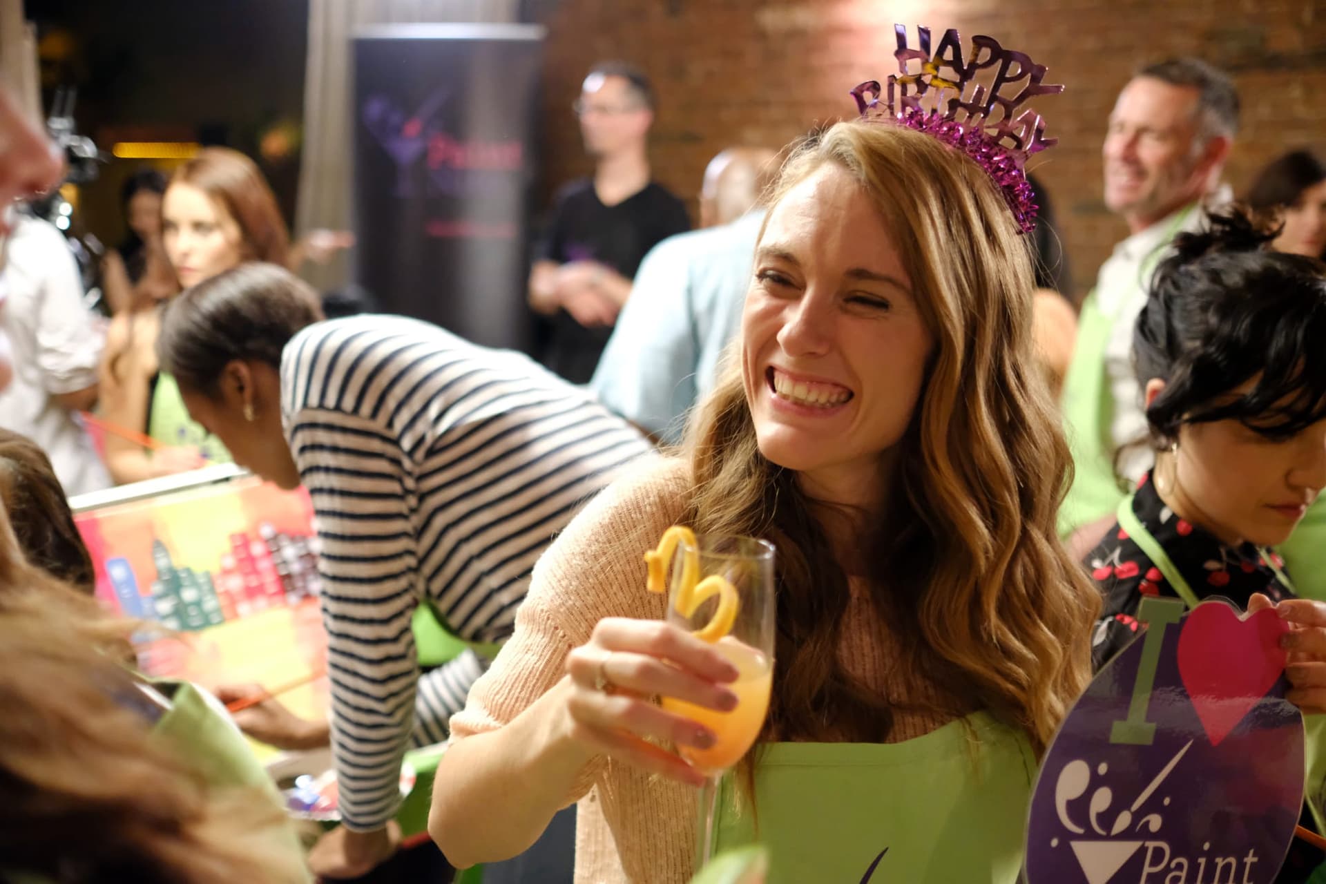 A guest in a Happy Birthday tiara laughing with a drink in hand at her private Paint Nite party, friends painting at easels around her