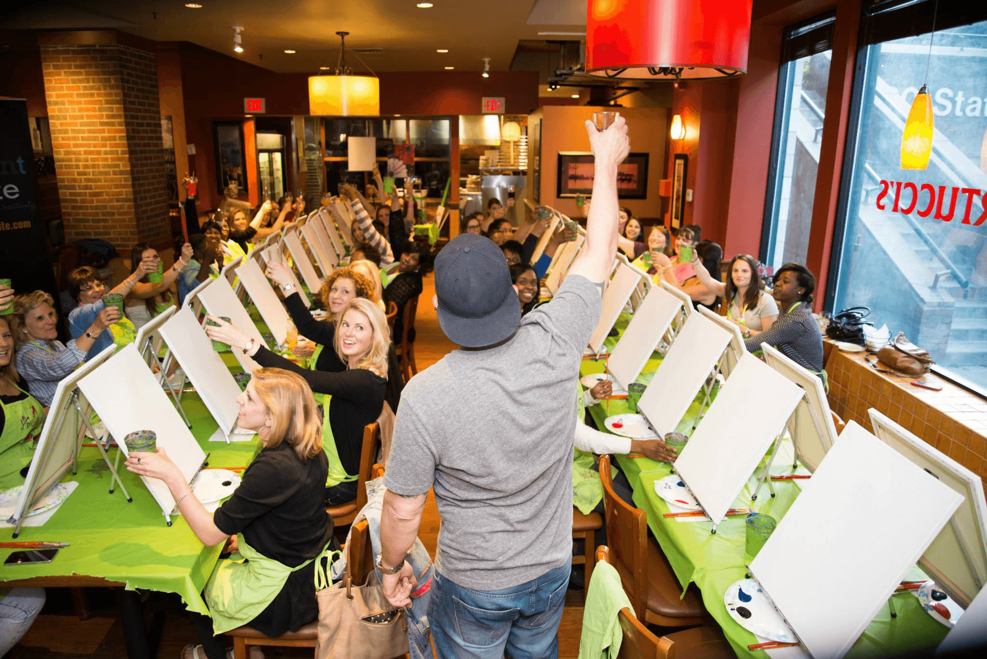 A Paint Nite host raising a glass to a full room of guests toasting back at their easels