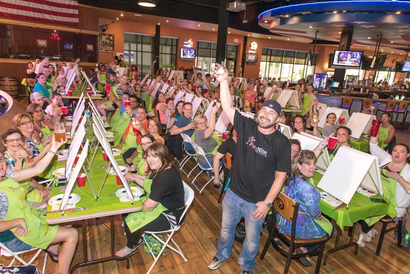 A packed venue of guests cheering at a Paint Nite paint and sip event